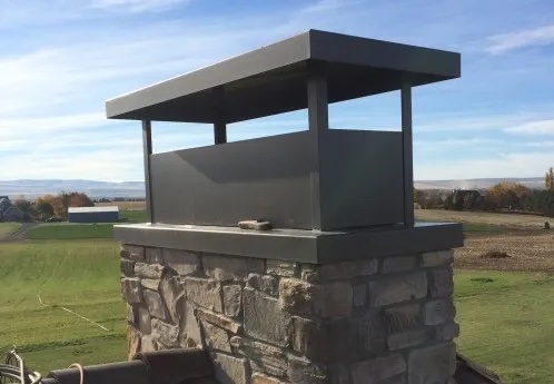 A stone chimney with a metal chimney cap, farmland and hills behind it