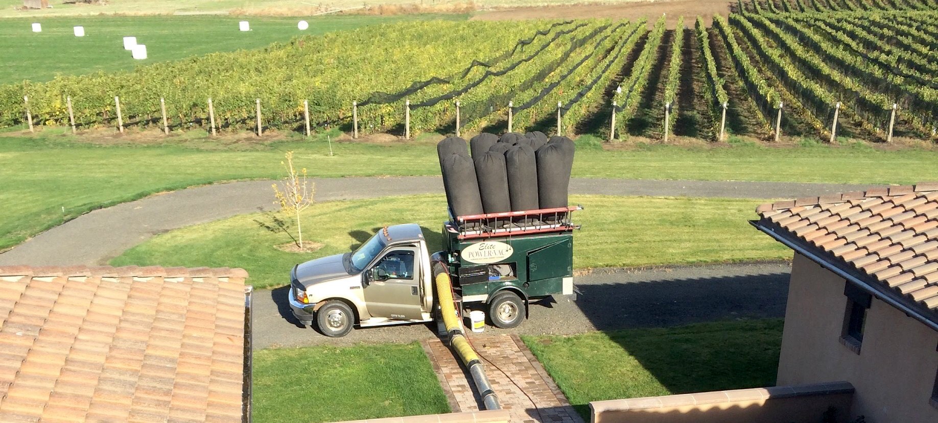 The Elite Power-Vac truck, its tall vacuum filter bags upright on the back, parked on a winery drive above rows of vineyard